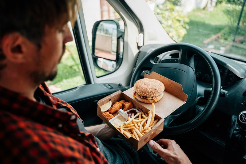 Man sitting in the driving seat of a car with a hamburger and friend on his lap.