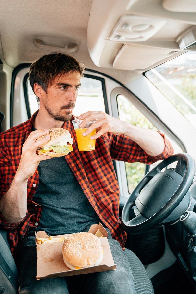 A man wearing a plaid red and black shirt sitting in his car drinking an orange juice and eating fast food.