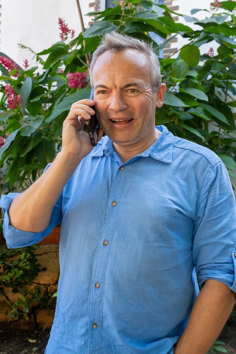 Smiling man talking on smartphone outdoors near green plants and flowers.