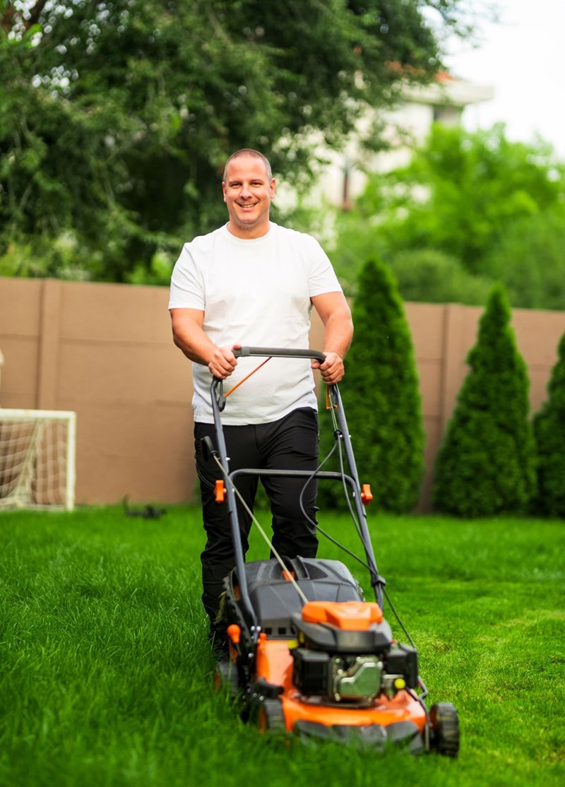 Man mowing lawn in backyard with push mower on green grass near trees and fence.