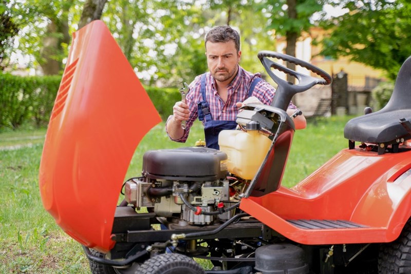 Man repairing riding lawn mower engine outdoors, holding wrench and checking parts on red mower in yard.