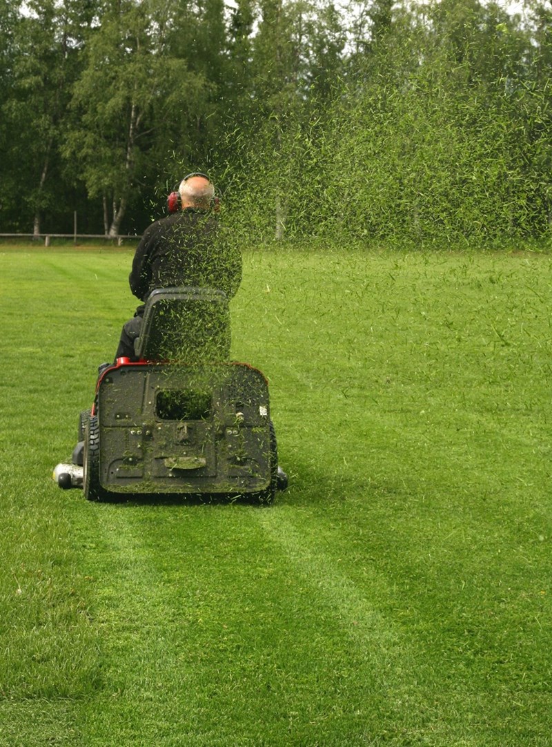 Man mowing lawn on riding mower, grass clippings spraying behind across a large green yard.