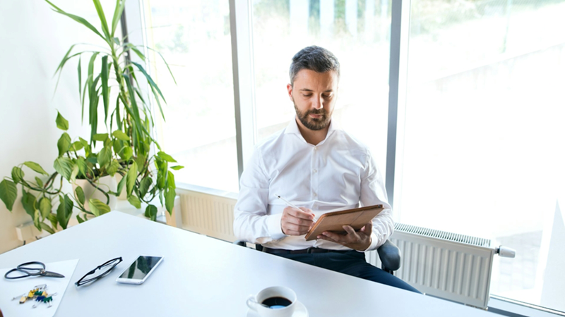 A man sits at a table and works on a tablet in an office with large glass windows