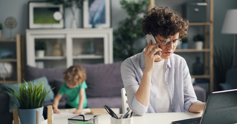 A woman sitting at a desk with a laptop talks on a cell phone with a child drawing on the coffee table behind her