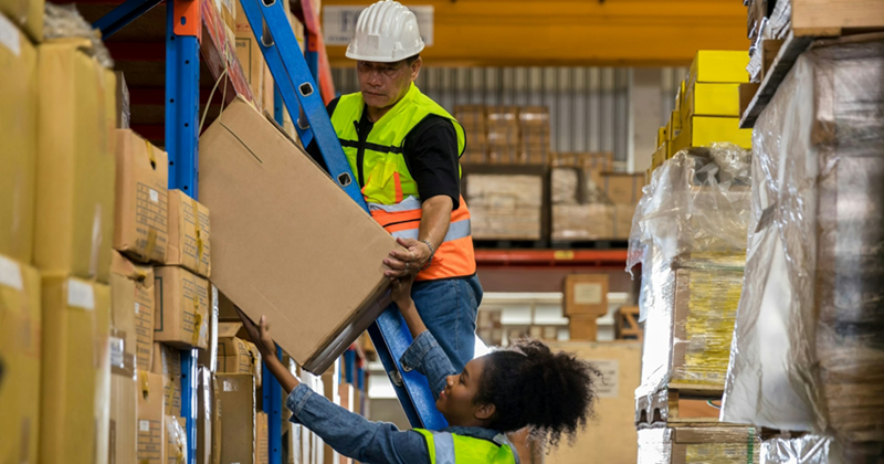 A warehouse worker standing on a ladder passes a box down to another worker standing on the ground