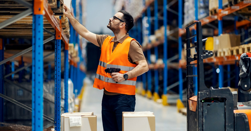 A warehouse employee reaches up to a pallet on a shelf in a warehouse