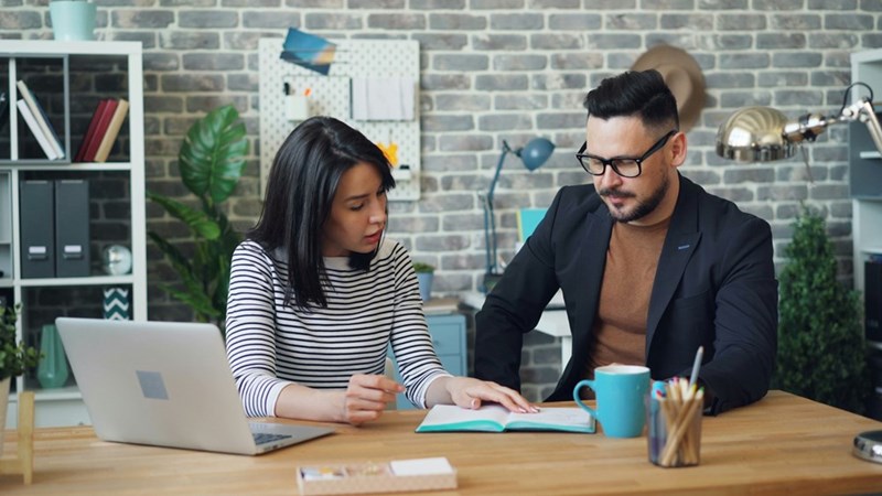 An employee takes a closer look at the notes her coworker made as the two sit down to work together.