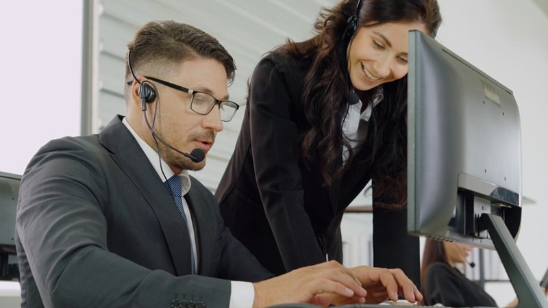 Call center supervisor assisting employee at desk, reviewing work on computer screen in office setting