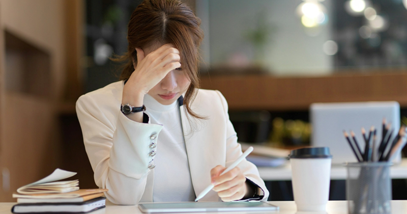 An employee sitting at a desk puts her hand to her forehead as she works on a tablet