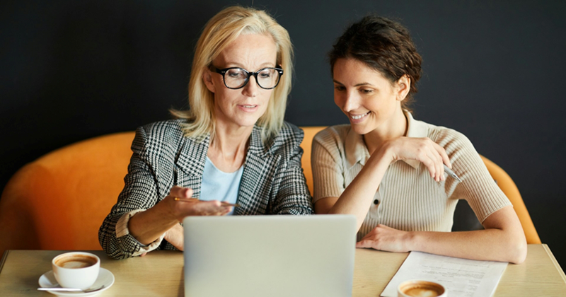 An older and younger woman sit together having coffee, discussing something on the laptop in front of them