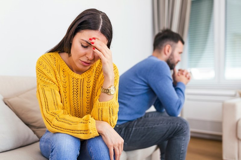 Woman wearing a yellow sweater is sitting on a couch ignoring her boyfriend.