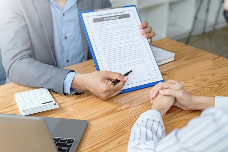 Man sitting on a desk, wearing a suit holds a contract and a pen signaling where to sign.