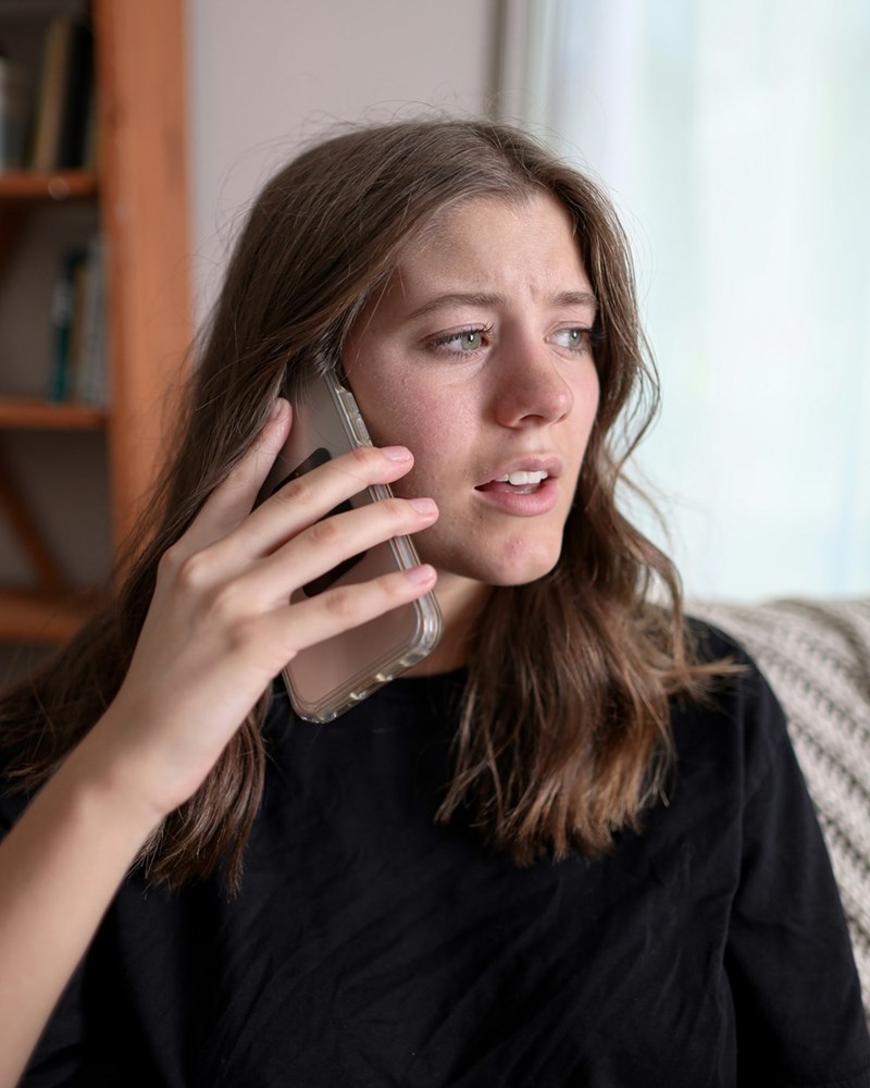 Woman sitting down on a couch wearing a black t-shirt talking over the phone looking concerned.