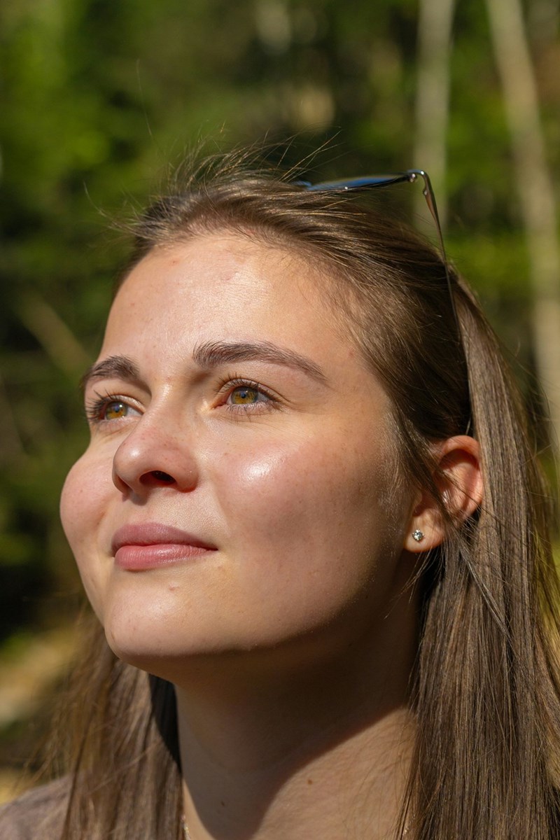 Close-up of young woman in sunlight looking up, glasses on head, soft forest background and warm natural light.