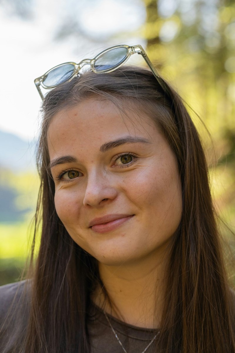 Close-up of smiling young woman outdoors with glasses on head, soft natural light and blurred green background.