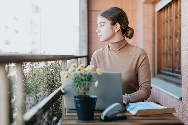 Woman using laptop on balcony, looking outside beside papers and plant in soft daylight.