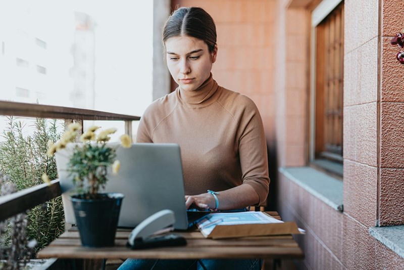 Woman typing on laptop on balcony table with papers and potted plant in soft daylight.