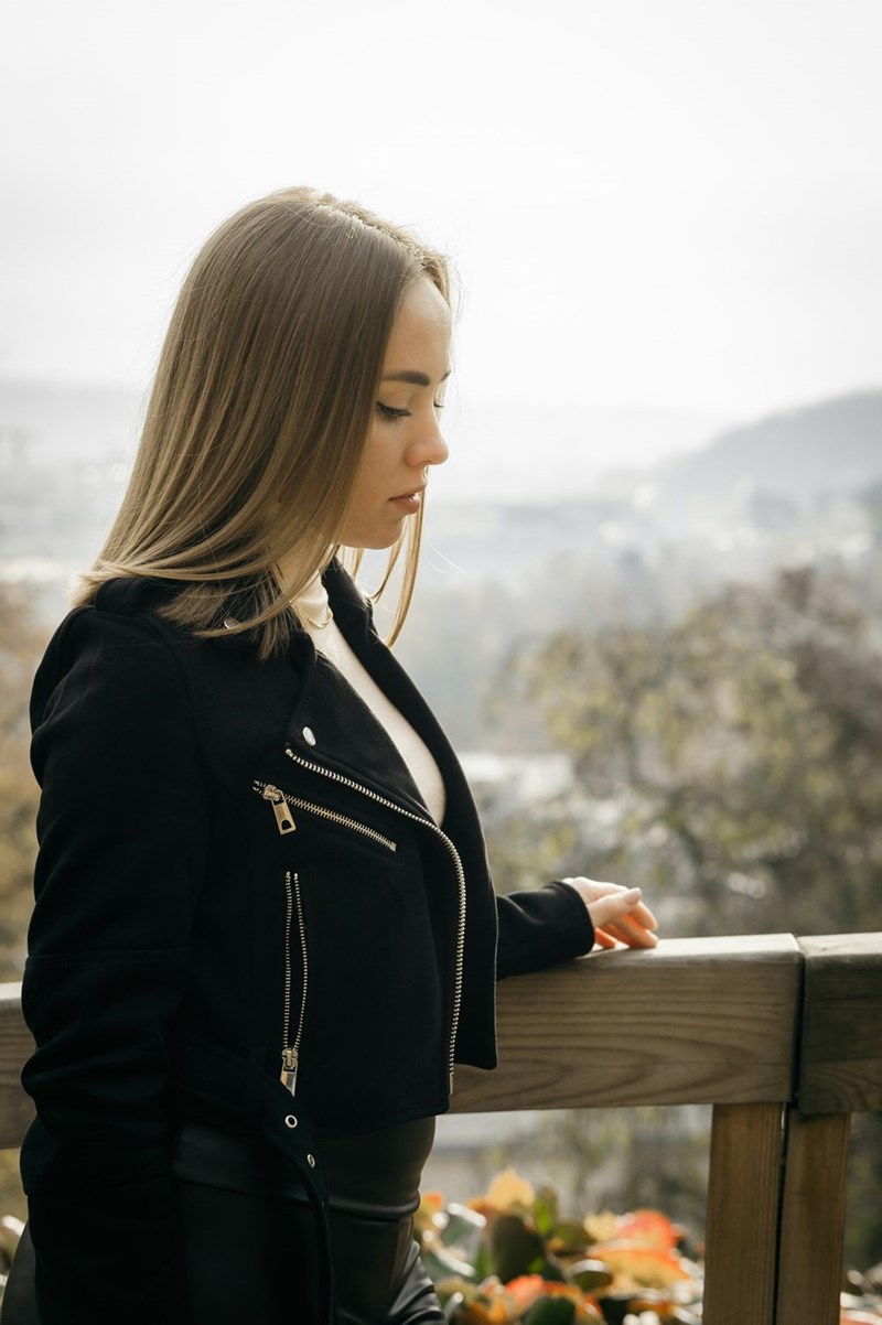 Woman in black jacket standing by wooden railing outdoors, looking down thoughtfully with blurred city in background.