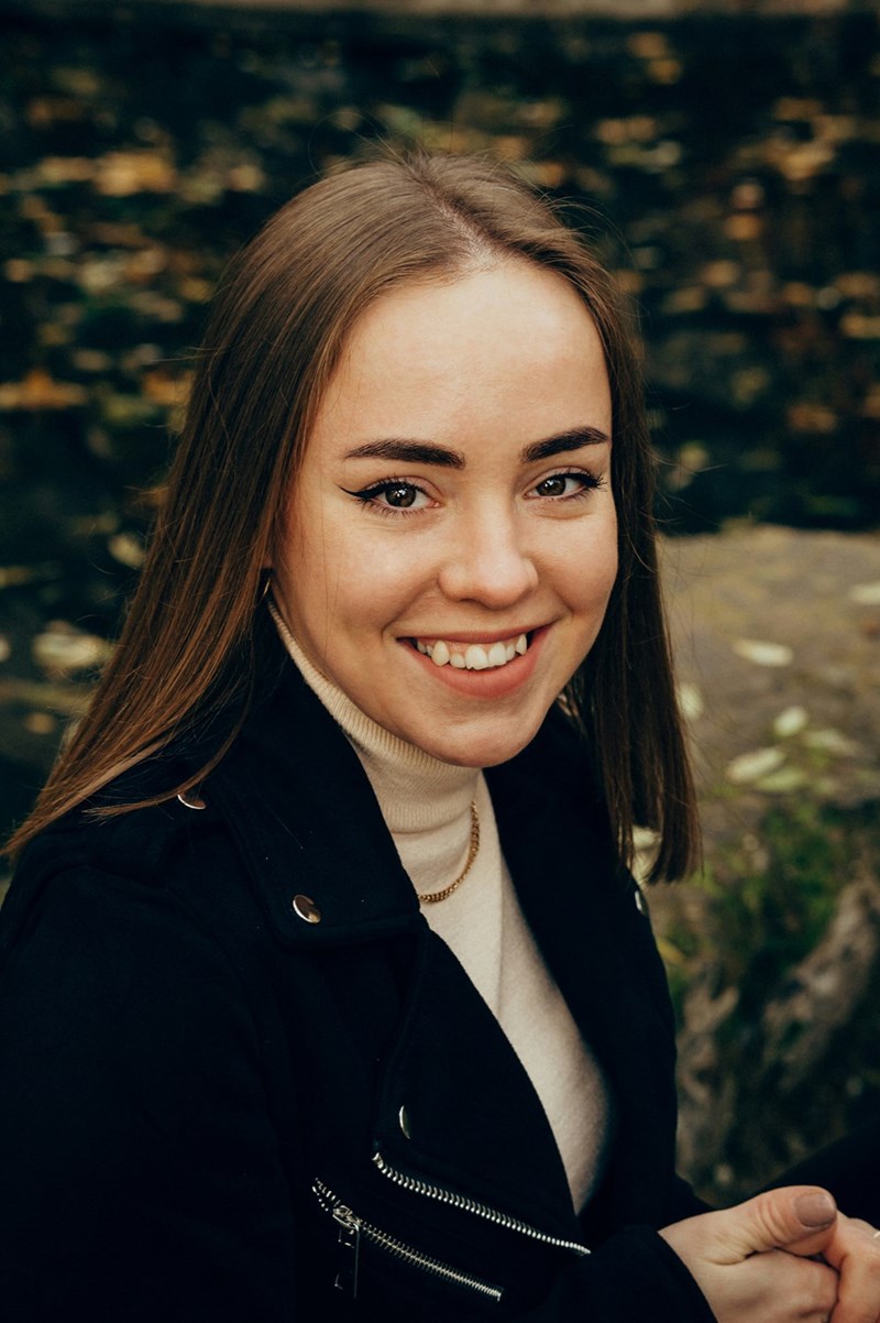 Smiling woman in black jacket outdoors near water, looking at camera with natural blurred background.