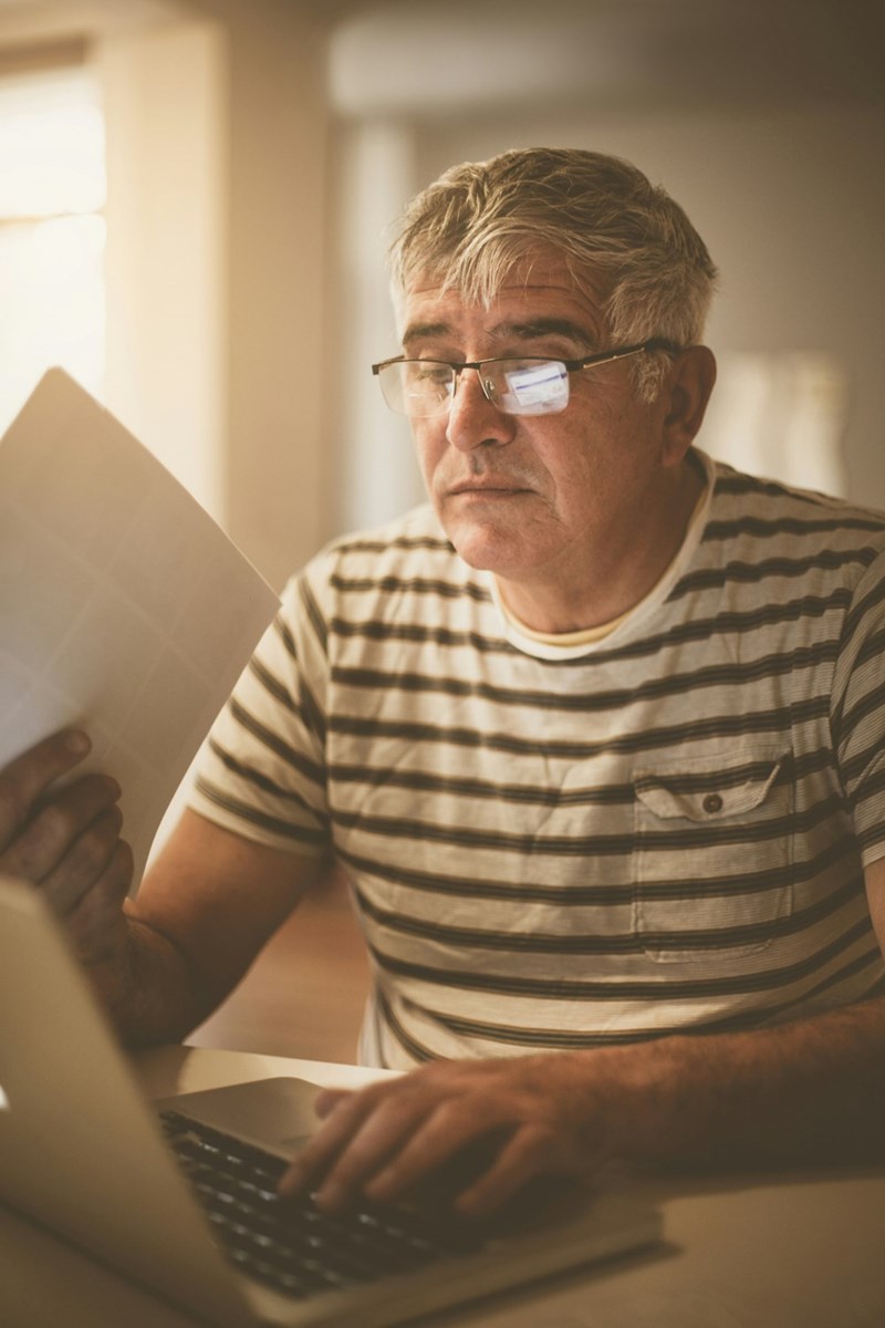 Older man with glasses reading document while using laptop at home in warm light, focused expression.