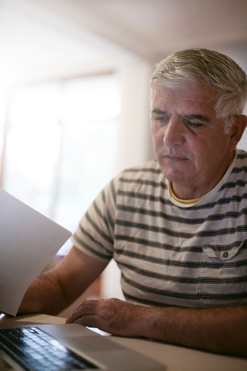 Older man reading document while using laptop at home, focused expression in soft natural light.
