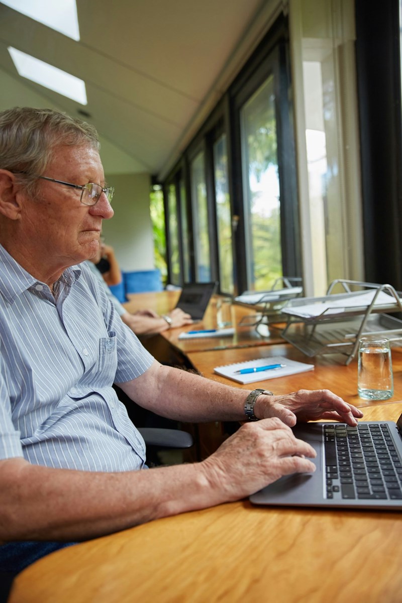 Older man typing on laptop at wooden desk by window in coworking space with natural light and glass of water.