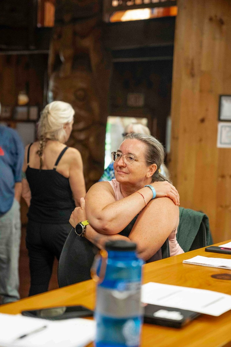 Woman with glasses seated at a wooden table, arms crossed over her shoulders, looking to the side in a casual indoor setting with people in the background.