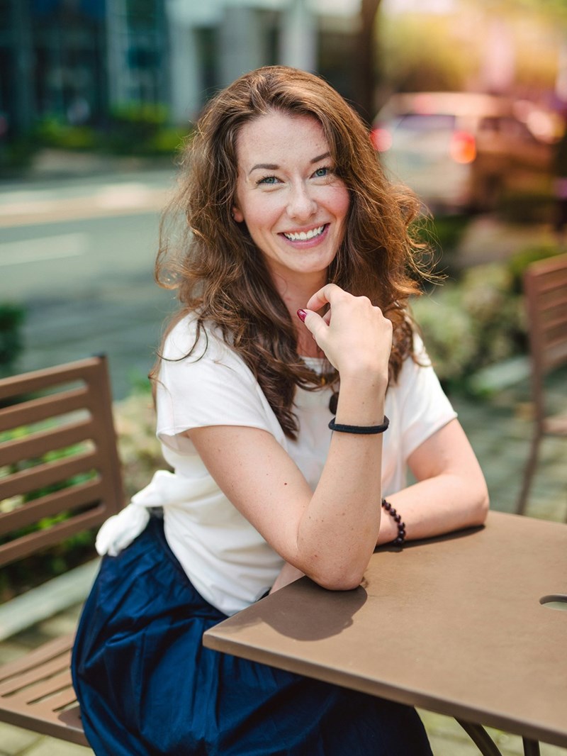 Smiling woman with curly hair sitting at an outdoor café table, resting her chin on her hand with a relaxed, friendly expression.
