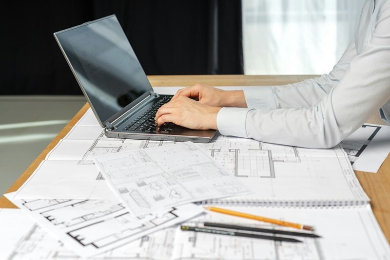 A professional uses a laptop over architectural plans, working on detailed designs.