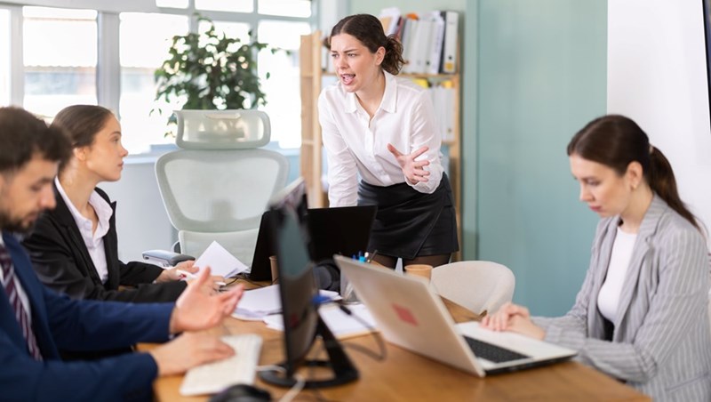 Woman standing and speaking emphatically during a tense office meeting while coworkers sit at a table with laptops and documents.