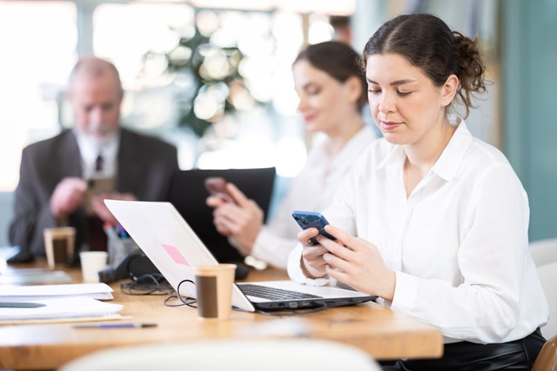 Woman checking her smartphone while sitting at a desk with a laptop, as coworkers behind her also look at their phones during a meeting.