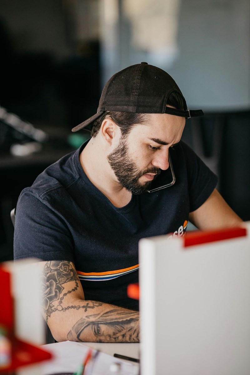 Man in a cap holding a phone between shoulder and ear while writing at a desk, focused on work with a laptop nearby.