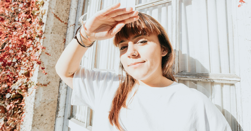A young woman shades her eyes from the sun as she stands outside, in front of a window with the curtains drawn and foliage around it