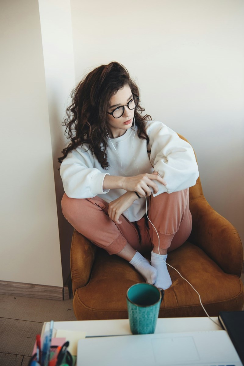 Curly-haired woman with glasses sits curled in chair, wearing earphones and adjusting sleeve in cozy indoor setting.