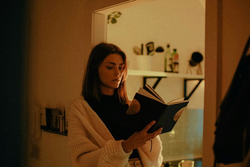 Woman reading a book in warm-lit kitchen, wearing cozy cardigan and standing indoors in soft evening light.