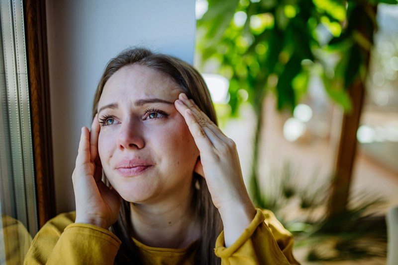 Emotional woman by window holds temples, teary-eyed and stressed in bright indoor setting with plants in background.