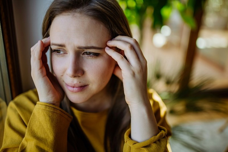 Close-up of stressed woman holding temples by window, looking worried and tense in bright indoor setting.