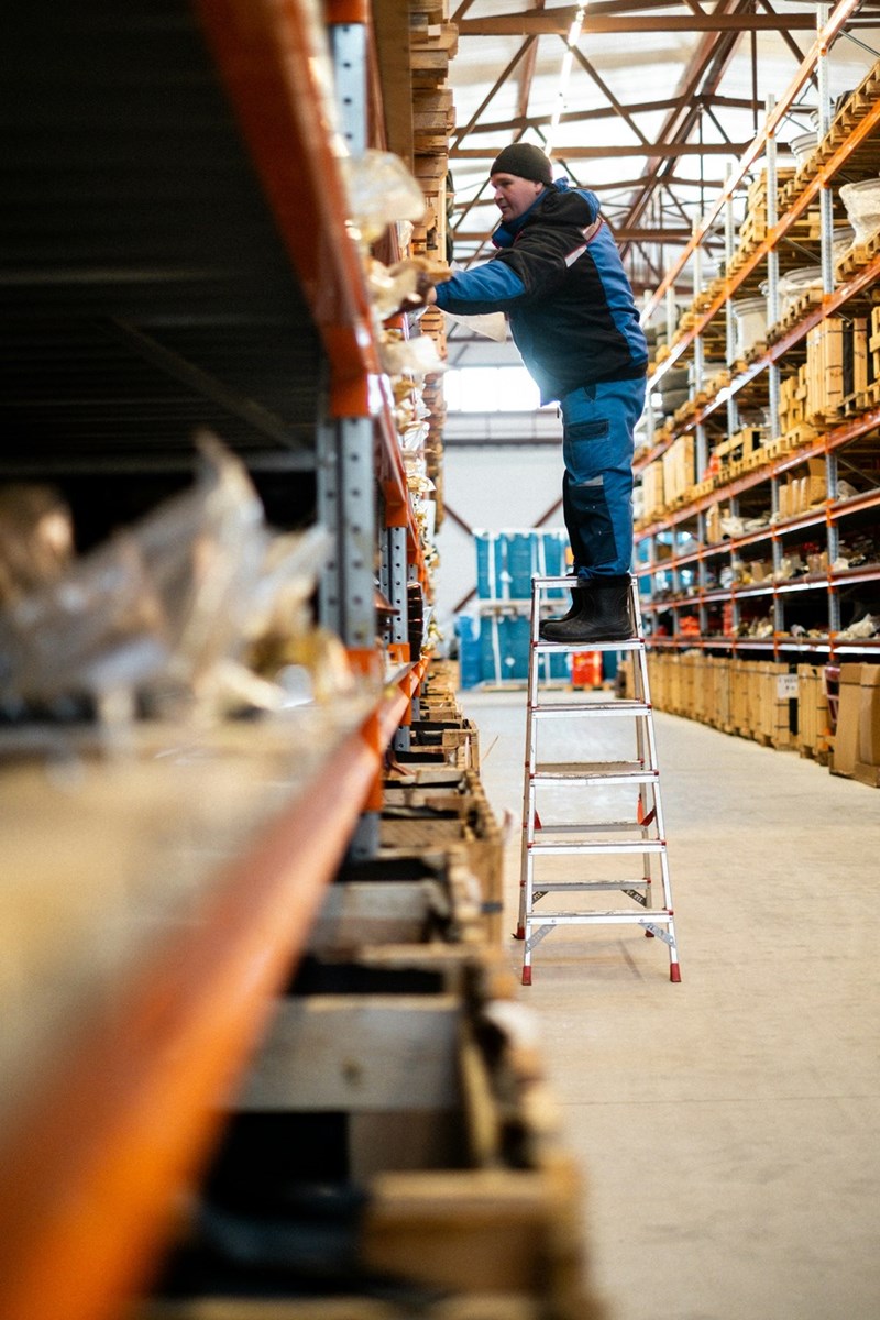 Hardware store employee stands high up in a ladder as he reaches for a product on a top shelf