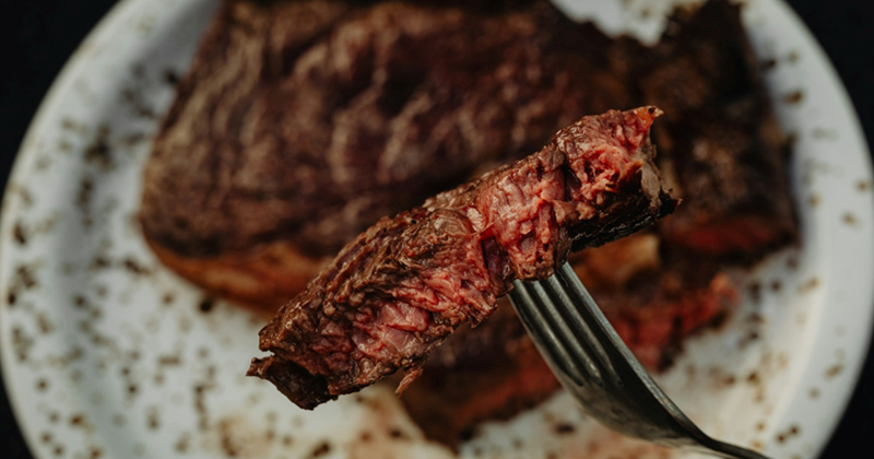 A fork holding a piece of steak above a plate with a medium-rare steak on it