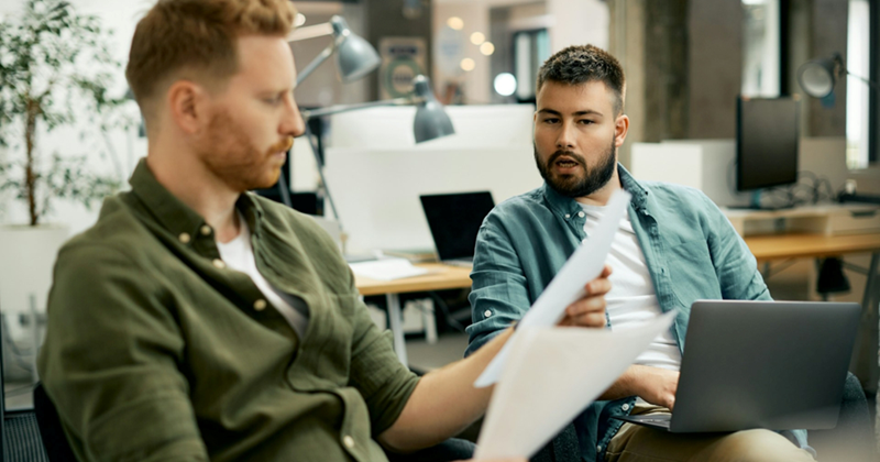 Two male coworkers sit together, one holding papers and the other a laptop as they have a discussion