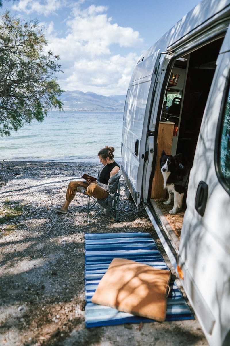 Woman and her dog relax beachside by their camper van.