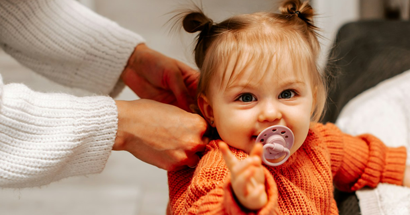 A caregiver adjusts the clothing of a female toddler with a pacifier as she leans against the couch