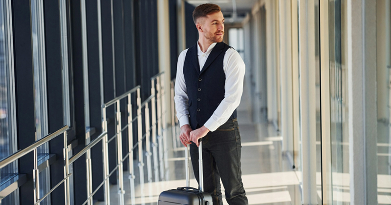 A young male professional looks out the window of an airport hallway, holding the handle of a small suitcase