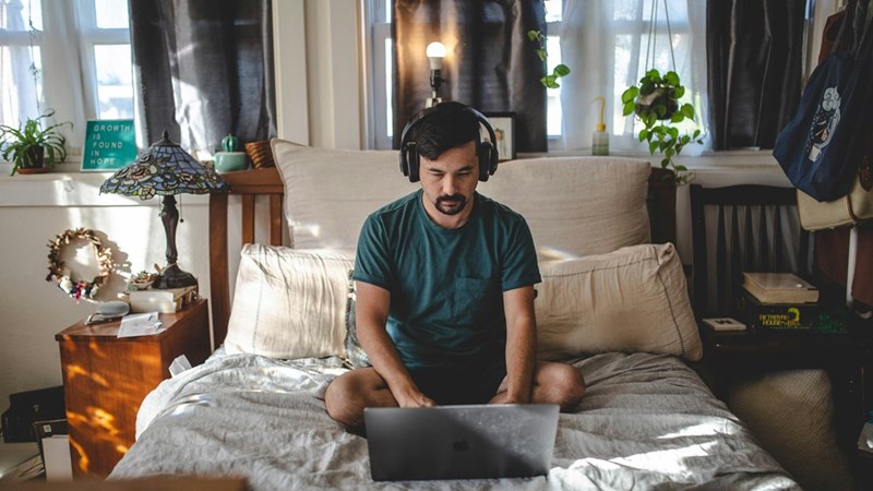 Man wearing headphones works on laptop while sitting cross-legged on bed in cozy sunlit bedroom.
