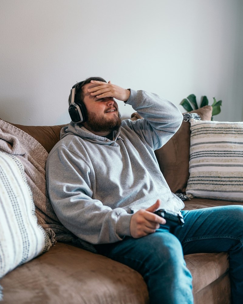 Frustrated gamer wearing headphones holds controller and covers face while sitting on living room couch.