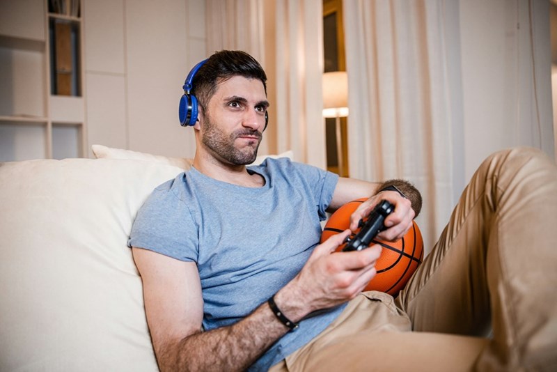 Man wearing headphones plays video games with controller while relaxing on couch at home.