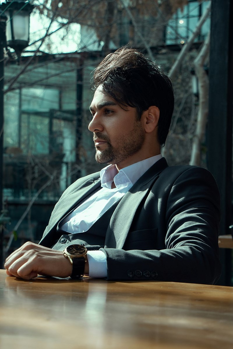 Businessman in black suit sitting outdoors at table with thoughtful expression.