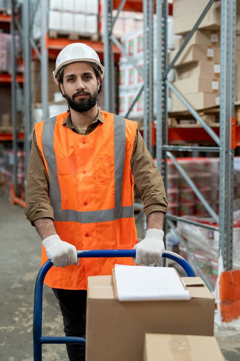 Content young man employee in reflective vest and hardhat working pushing storage cart over warehouse