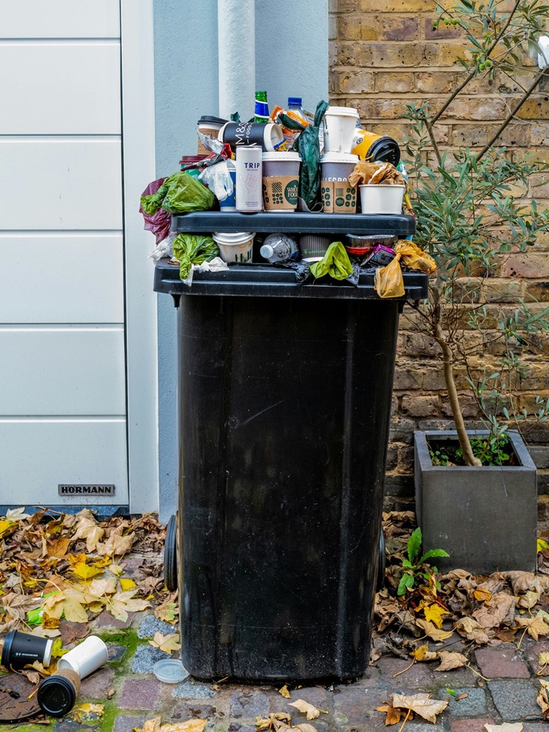 A black garbage can by the side of a house overflowing with trash.