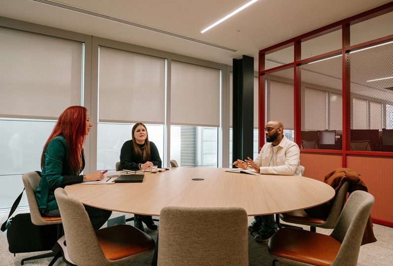 Three coworkers sit in a conference room around a circular table in their office.
